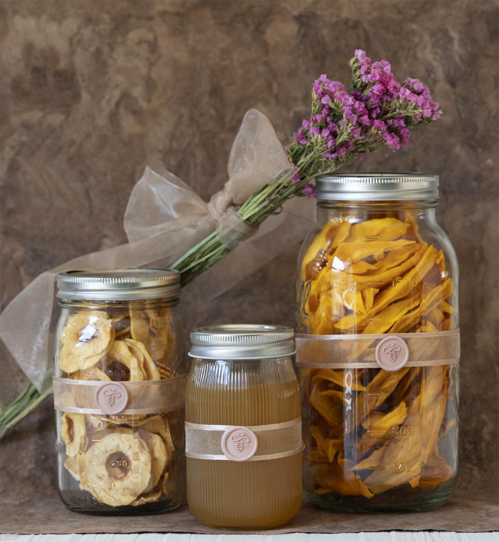Glass jars with dried fruit and honey inside, decorated with a beige ribbon and a sealing medallion with a champagne-colored bee figure.