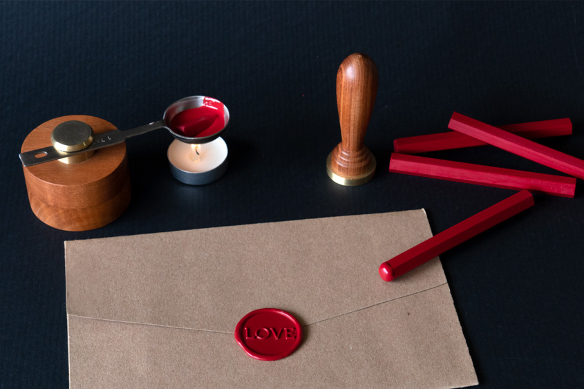 Kraft paper envelope that has been sealed with the word LOVE in red wax. There is a small wax melter with a lighted candle and some sticks of red wax