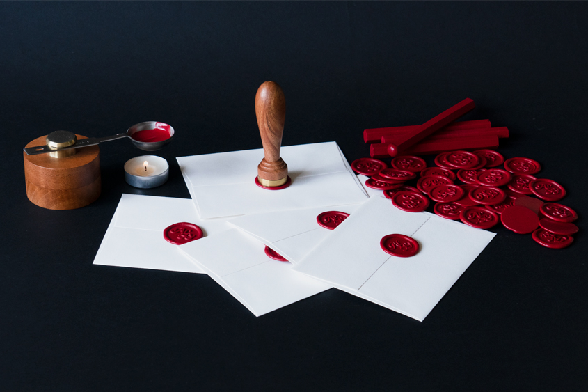 Mini melting base that holds a stainless steel spoon in which red sealing wax is melted. Some white envelopes have been sealed with the initials ALB i