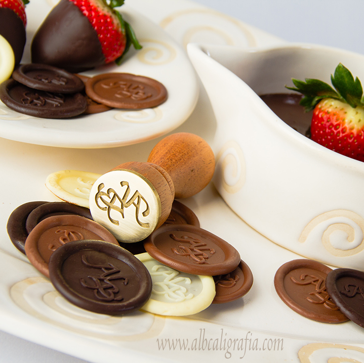 Metallic seal to seal chocolates, surrounded by chocolates stamped with the seal of ALB and some strawberries covered with chocolate on white ceramic 